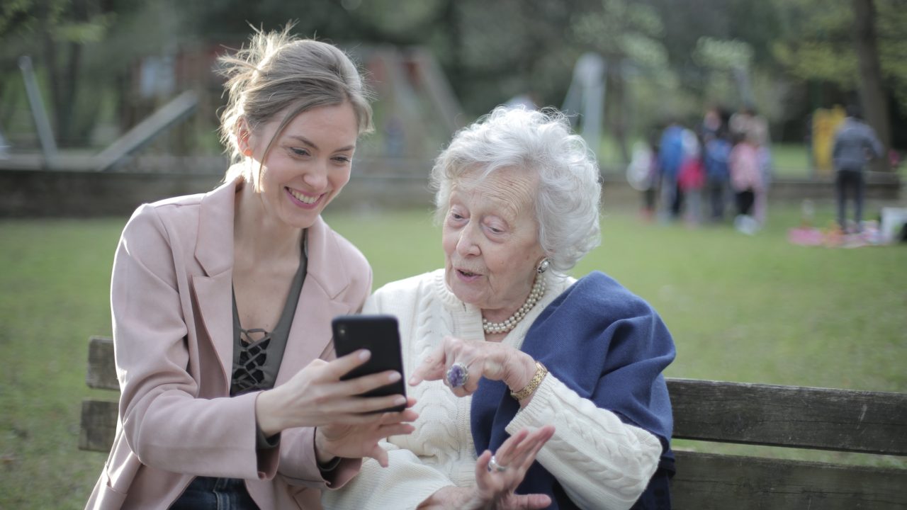 caring for aging parents. A Women Caring For Her Mother As She Teaches Her Something New On Her Cell Phone At The Park
