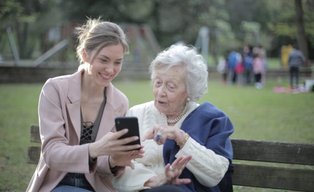 A Women Caring For Her Mother As She Teaches Her Something New On Her Cell Phone At The Park